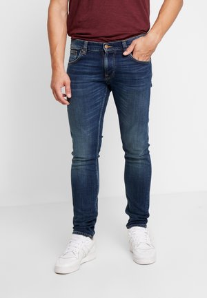 Homme portant un jean slim bleu foncé, des baskets blanches et un t-shirt rouge foncé, debout avec une main dans la poche.