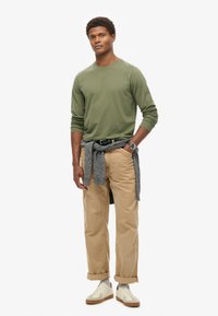 Camisa de manga larga verde oliva, pantalones caqui con los puños enrollados, suéter gris atado a la cintura y zapatillas blancas con suelas beige.