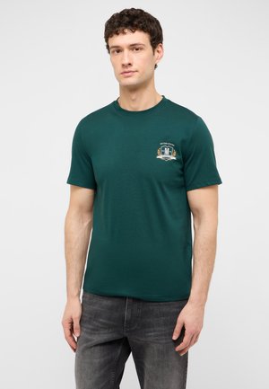 Young man wearing a dark green short-sleeve t-shirt with a small emblem on the chest and dark gray jeans standing against a plain background.