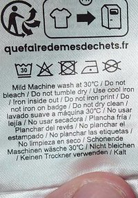 Vård- och tvättråd med tvättinstruktioner, tryckt text, ikoner för tvätt, torkning och strykning, främst vit tyg med svart text.