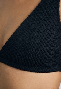 Czarny dzianinowy biustonosz bikini o teksturowanej tkaninie, trójkątnym kształcie i elastycznym pasku. Zbliżenie ukazujące szczegóły szycia.