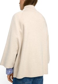 Beżowy, oversize sweter z wysokim kołnierzem, miękką teksturą i rękawami raglanowymi. Nosi się go na paskowanym niebieskim tle i zestawia z niebieskimi dżinsami.