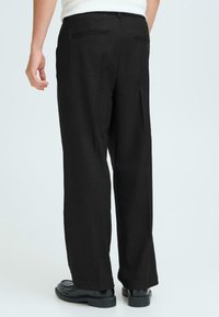Pantalons larges noirs avec une texture lisse, comportant deux poches arrière et une taille épurée, associés à des chaussures noires brillantes.