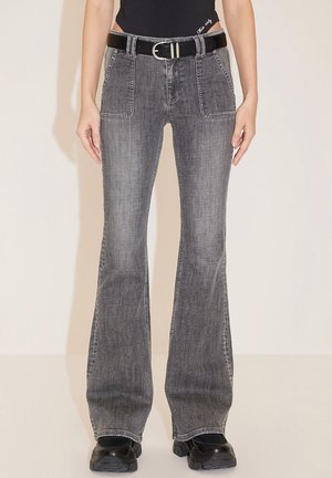 Kobieta nosząca wyblakłe czarne dzwoniaste dżinsy z dużymi przednimi kieszeniami, czarny pasek, czarne buty i czarną bluzkę na jednolitym tle.