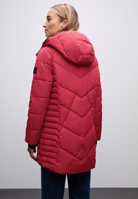 Abrigo acolchado rojo con capucha, que presenta costuras en forma de chevrones, una superficie texturizada y bolsillos laterales. El diseño está hecho para un ajuste ceñido.