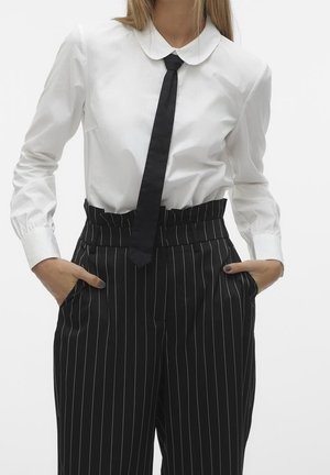 Vrouw draagt een wit overhemd met lange mouwen en een Peter Pan-kraag, een smalle zwarte das en een hoog getailleerde zwarte pantalon met krijtstreep, handen in de zakken.