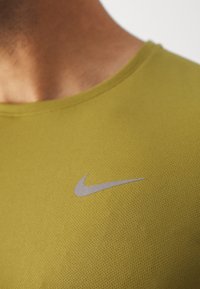 Camiseta deportiva verde con cuello redondo, que presenta una sutil tela texturizada y un pequeño logotipo plateado de Nike situado en el pecho.