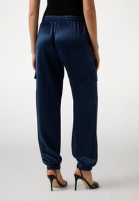 Marinblå satinjoggers med elastisk midjeband, elastiska muddar och sidoficka. Slät yta, avslappnad passform och minimalistisk design.