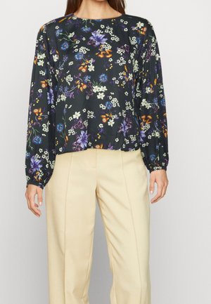 Femme portant un chemisier noir manches longues à fleurs violettes, blanches et oranges, associé à un pantalon beige sur un fond clair.