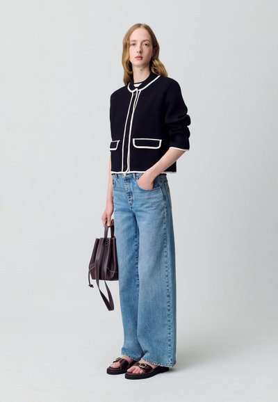 Cardigan nero a maniche corte con piping bianco, abbinato a jeans larghi azzurri. Il modello tiene una borsa a secchiello marrone scuro e indossa sandali piatti.