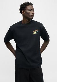 Άνδρας που φοράει μαύρο μπλουζάκι New Balance Running, στέκεται με τα χέρια πίσω από την πλάτη, κοιτάζοντας προς τα πλάγια σε απλό φωτεινό φόντο.