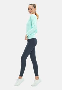 Langärmliges türkisfarbenes Oberteil, figurbetonte schwarze Leggings, weiße Sneakers. Glatter Stoff, taillierter Schnitt, minimales Logo-Detail auf den Leggings.