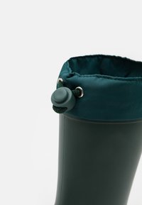 Botte en caoutchouc vert foncé avec une surface texturée, dotée d'un haut côtelé rembourré en bleu sarcelle et d'un cordon de serrage pour l'ajustement.