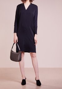 Tummansininen mekko, jossa on V-pääntie ja pitkät hihat, varustettuna nappikoristeella. Yhdistettynä pieneen harmaaseen käsilaukkuun ja mustiin liukuviin kenkiin.