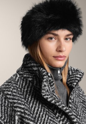 Črna kapa iz umetnega krzna v kombinaciji z teksturiranim, vzorčastim sivim plaščem. Viden je visok ovratnik in detajl z zadrgo na sivem puloverju z visokim ovratnikom spodaj.