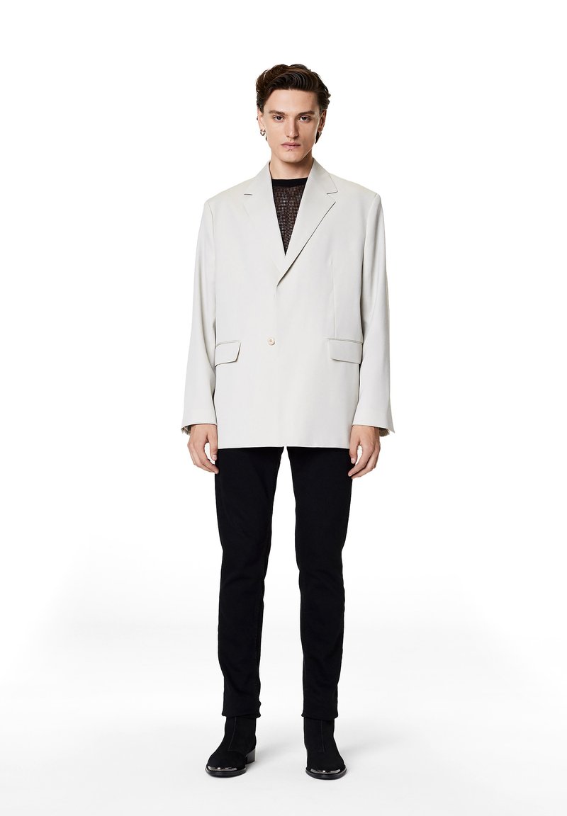 Light beige oversized blazer with notch lapels, single button closure, two front pockets, worn over a black mesh top and slim black pants.