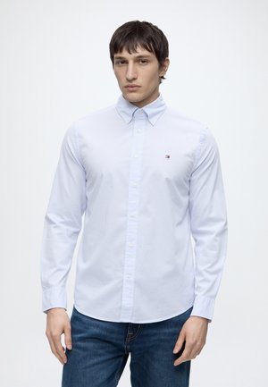 Homme portant une chemise à carreaux bleu clair boutonnée avec un petit logo sur la poitrine et un jean bleu foncé, debout devant un fond uni.
