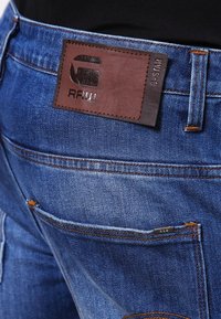 Blå denimjeans har en läderetikett med präglad branding, kontrasterande sömmar och två bakfickor. Detaljen inkluderar en liten logotypetikett.