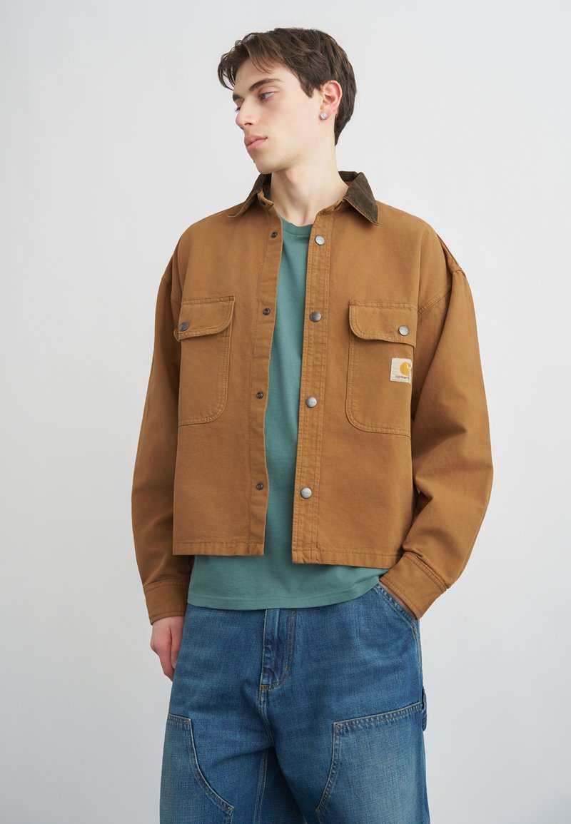 Junger Mann, der eine braune Jacke mit Knöpfen über einem türkisfarbenen Hemd und lockeren blauen Jeans trägt, blickt nach links mit einer Hand in der Tasche.