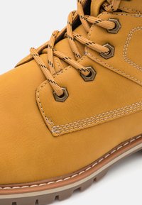 Botas de cuero marrón con ojales hexagonales, cordones en tono beige y acentos de costura en contraste. La suela resistente ofrece tracción y durabilidad.