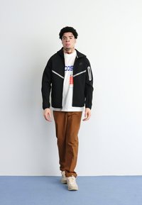 Jacket preta com fecho éclair e detalhes cinzas, t-shirt branca com texto azul, calças castanhas e sapatilhas claras; em pé contra um fundo simples.