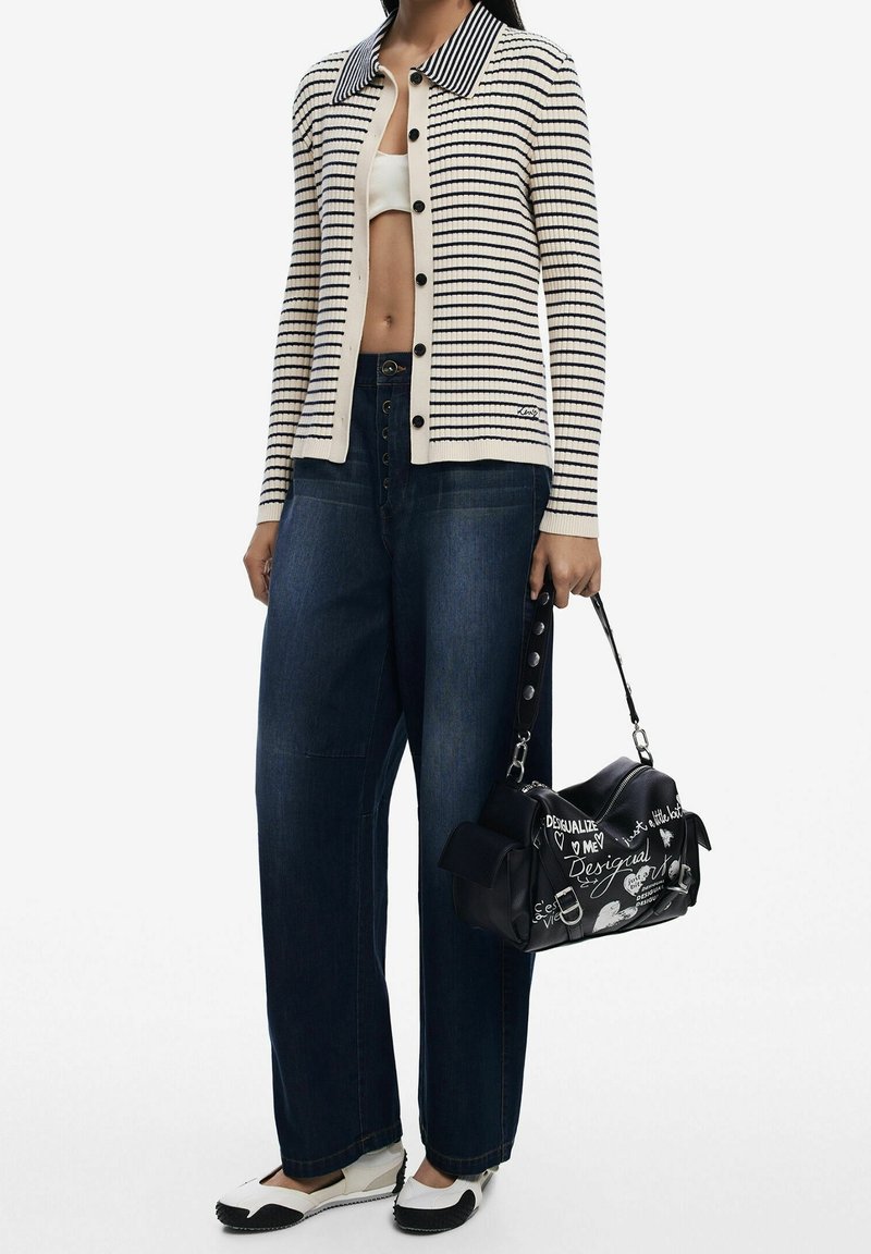 Femme portant un cardigan rayé crème et noir, un crop top blanc, un jean large foncé, des chaussures blanches et noires, tenant un sac à main noir avec des imprimés blancs.
