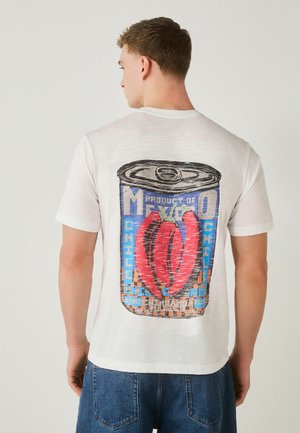 Muž v bielom tričku s veľkou grafikou mexického chilli v červenej, modrej a čiernej farbe na chrbte, v kombinácii s modrými džínsy.