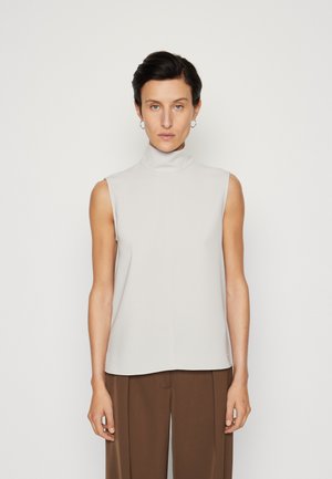 Sleeveless high-neck top in light gray fabric, featuring a seamless, smooth texture and minimalistic design, paired with brown trousers.