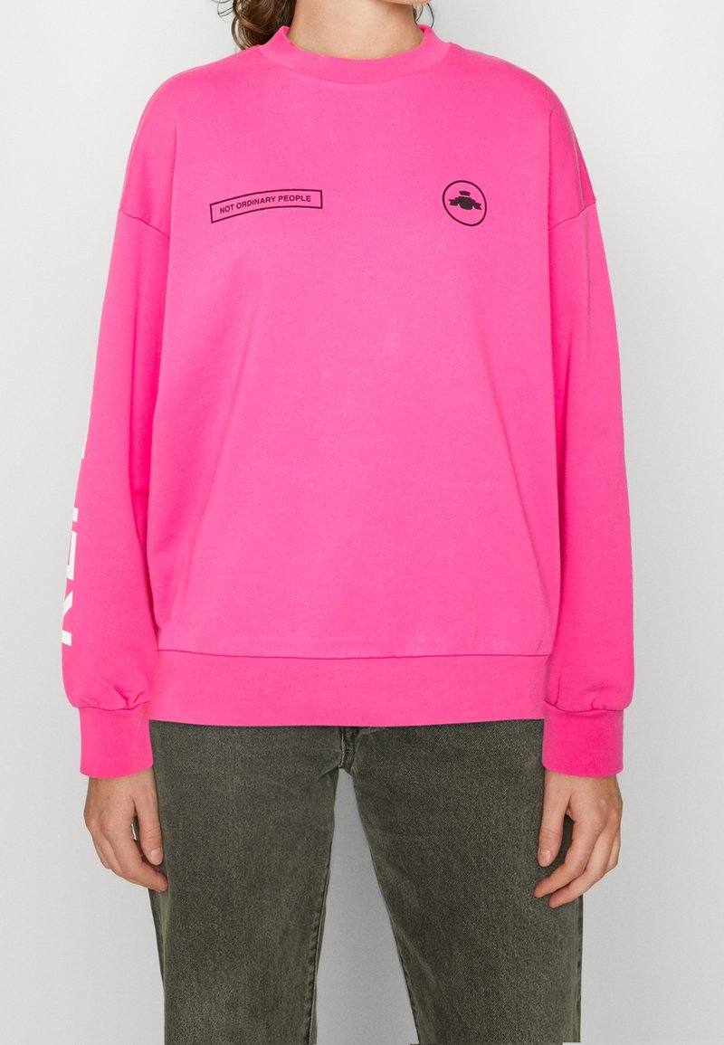 Ljusröd sweatshirt med rund hals, sänkta axlar och långa ärmar. Har svart text och en cirkulär grafik på bröstet.