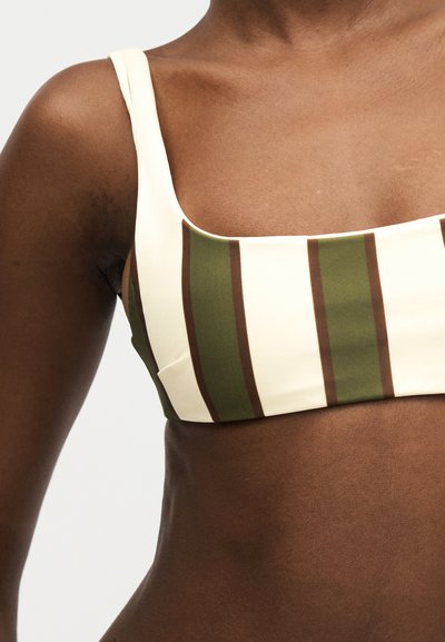 Γυναίκα με κορυφή εκρού χρώματος με κατακόρυφες φαρδιές ελιά πράσινες ρίγες και λεπτά καφέ περιγράμματα, που δείχνει τον άνω κορμό και τον ώμο της.