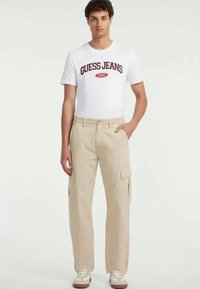 Bežid прациташ pants, millel on küljetaskud ja mugav lõige, koos valge T-särgiga, millel on punane ja must logo.