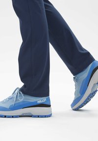 Baskets de sport bleu clair avec une tige texturée, des accents gris et une semelle sombre ; portées avec un pantalon marine. Présente le logo de la marque sur le côté.