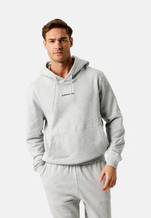 Mees kannab helehallist kapuutsiga pusa ja sobivaid dressipükse, mille rinnal on kiri "BJÖRN BORG"; seisab ühe käega taskus.