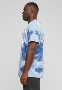 Modré tričko z bavlny s krátkým rukávem a technikou tie-dye, s kulatým výstřihem a grafickým potiskem na zadní straně. Kombinováno s tmavými džíny.