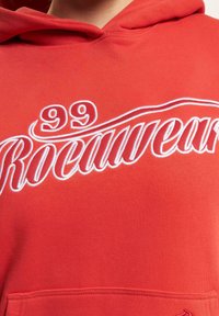 Rød hettegenser med hvit brodert tekst "99 Rocawear" og en glatt tekstur, samt frontlomme. Hette har et klassisk design.