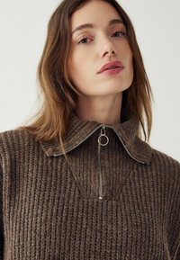 Brūns adīts džemperis ar ribotu tekstūru, augstu apkakli un pusi sānu rāvējslēdzēju, kurā ir apaļš sudraba pievilkšanas elements. Tīrs, minimālisma dizains.