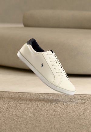 Fehér alacsony szárú tornacipő fekete belsőrésszel és fűzős elülső résszel, oldalán egy kis fekete madár logóval, bézs texturált felületű háttéren.