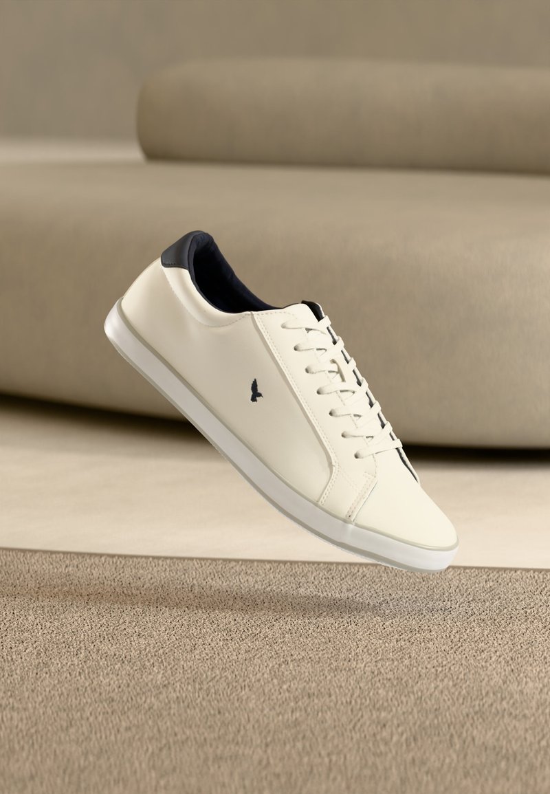 Sneaker bianco low-top con interno nero e chiusura frontale con lacci, con piccolo logo di un uccello nero sul lato, su sfondo beige con superficie strutturata.