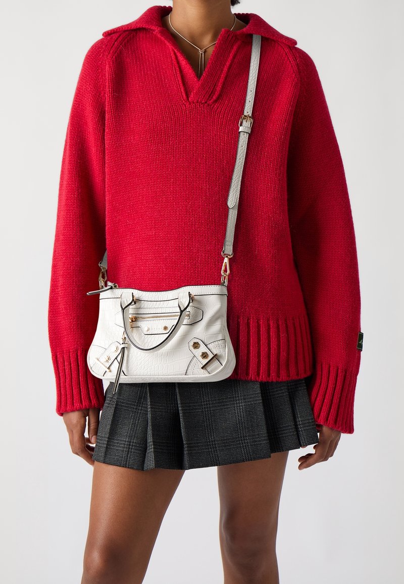 Personne portant un pull rouge en maille et une jupe plissée noire, tenant un sac bandoulière blanc avec des détails en métal doré.