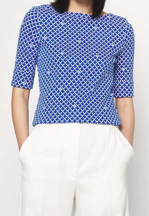 Femme portant un haut à manches courtes bleu à motifs, rentré dans un pantalon blanc taille haute, les mains dans les poches.