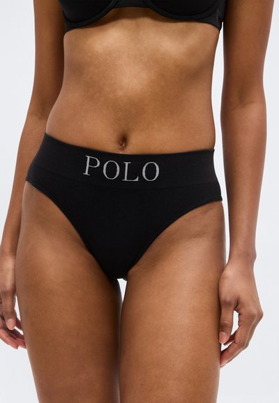 Femme portant un slip taille haute noir avec le logo "POLO" sur la ceinture et un soutien-gorge noir assorti, vue de la moitié du torse jusqu'aux cuisses.