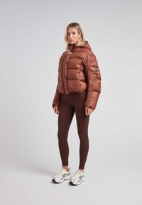 Brun puffjakke med et cropped design, store quiltede sektioner, frontknapper og hætte. Sammen med brune leggings og hvide sneakers.