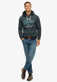 Grijze hoodie met witte print, blauwe jeans en bruin veterlaarzen. De hoodie heeft een trekkoord en textured stof. Casual outfit.