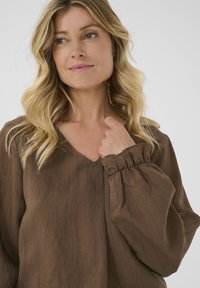 Blond kvinna med vågigt hår, iklädd en brun långärmad blus med samlade ärmslut, tittande något åt sidan med ett milt leende.