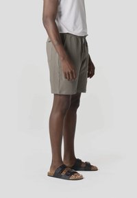 Hombre vestido con pantalones cortos texturizados color topo, camisa blanca y sandalias negras de dos tiras, de pie contra un fondo claro y liso.