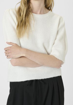 Person with long hair wearing a white short-sleeve sweater and black pants, arms crossed over the chest, neutral background.
