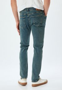 Jeans in denim blu sbiadito con taglio dritto, dotati di due tasche posteriori e una toppa in pelle, abbinati a sneakers bianche con suole marroni.