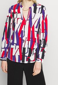 Vit blus med abstrakt mönster i rött, lila och svart. Har en henley-krage, knapplist och långa ärmar. Slät textur.