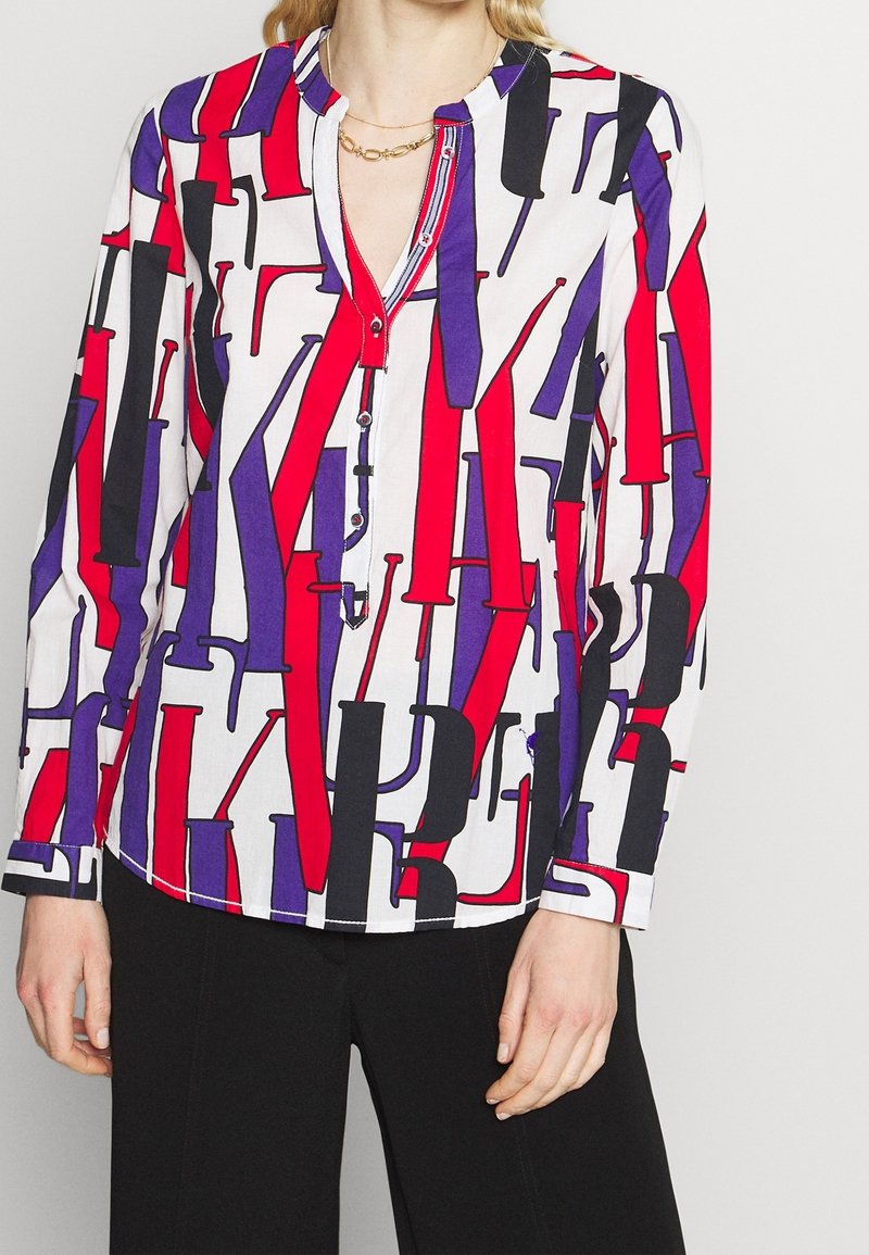 Vit blus med abstrakt mönster i rött, lila och svart. Har en henley-krage, knapplist och långa ärmar. Slät textur.