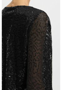 Haut noir transparent avec un motif texturé orné de sequins. Manches longues avec une finition lisse, mettant en valeur un design ajusté.